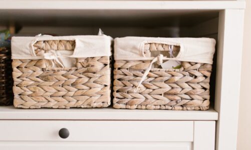 Dwa plecione kosze z jasnymi materiałowymi wkładami na białej półce mebla.