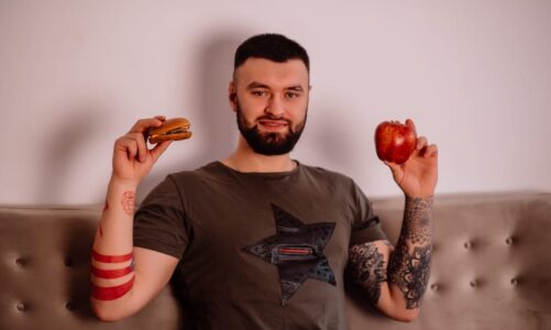 Mężczyzna z tatuażami trzyma w dłoniach hamburgera oraz czerwone jabłko.