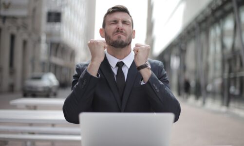 Mężczyzna w garniturze siedzi z zaciśniętymi pięściami przed laptopem na zewnątrz.